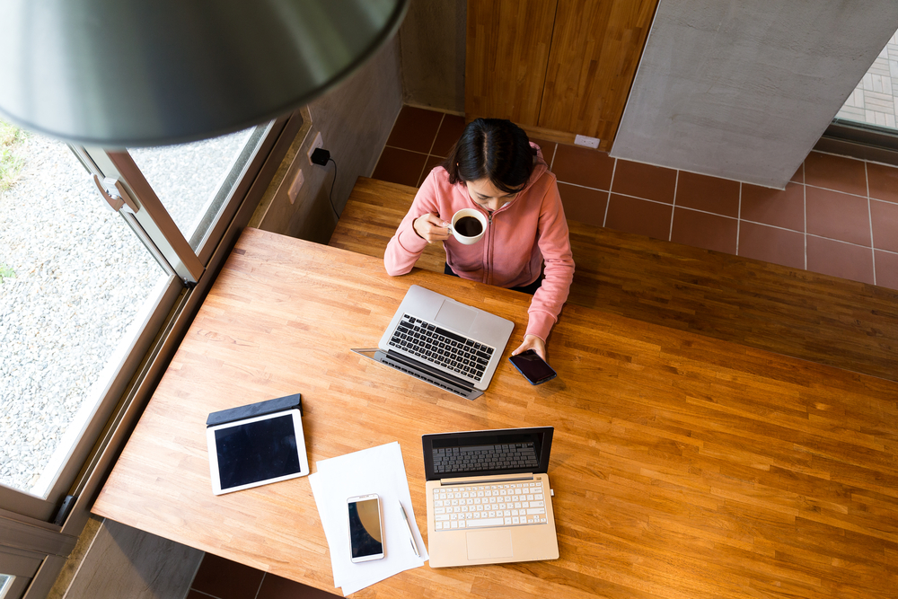 Woman drinking coffee checking message on cell phone with laptop and papers in front of her